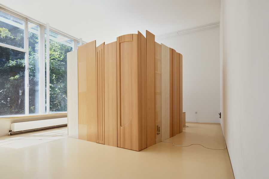  Installation view of Charms (2025) by Julia Rosenstock: a 30-minute video projected within a custom frame, accompanied by two-channel sound transmitted through resonant surfaces in the gallery. A bench sits in the space alongside wall-mounted reliefs made from plastic foil-coated MDF with walnut and oak veneer.