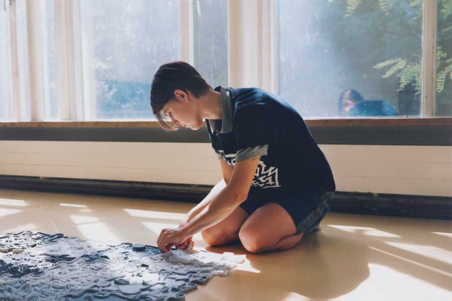 Photo of a bright room with sunlight streaming through large windows onto a yellow floor covered in silicone tiles arranged like a puzzle. The artist Julia Rosenstock is sitting next to the installation, lifting one tile while looking down at the work. The tiles are part of the 2024 work "Love story of a Ghost".