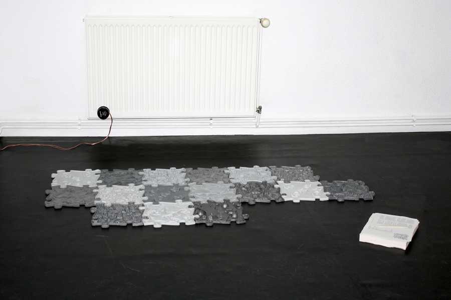 A flash-lit installation view of the work "Love story of a ghost" by Julia Rosenstock, installed at New Fears Gallery, Berlin, 2024. A transducer is attached to a heater, silicone mats are lying on the black floor in front of it. The mats have interlocking puzzle-like edges and vary in shades of gray. On the right side, a stack of paper on top of it lays a folding bone. 