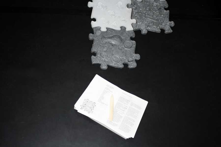 A flash-lit detail view of the work "Love story of a ghost" by Julia Rosenstock, installed at New Fears Gallery, Berlin, 2024. A silicone tile in the shape of a puzzle photographed from above.Underneath a stack of paper on top of it lays a folding bone. 