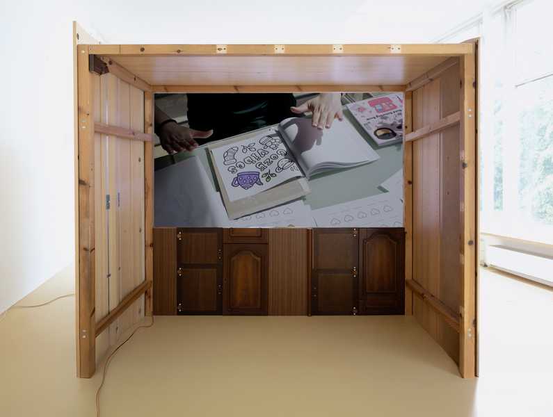  Installation view of Charms (2025) by Julia Rosenstock: a 30-minute video projected within a custom frame, accompanied by two-channel sound transmitted through resonant surfaces in the gallery. A bench sits in the space alongside wall-mounted reliefs made from plastic foil-coated MDF with walnut and oak veneer.